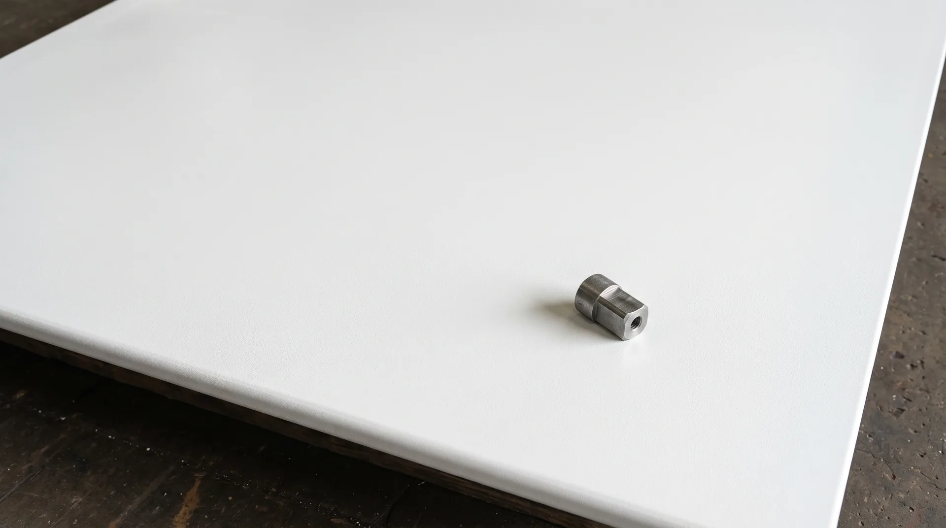 A large plain white panel lying flat and filling the frame, one small dull grey machined metal part resting near a corner, bench showing along one edge.