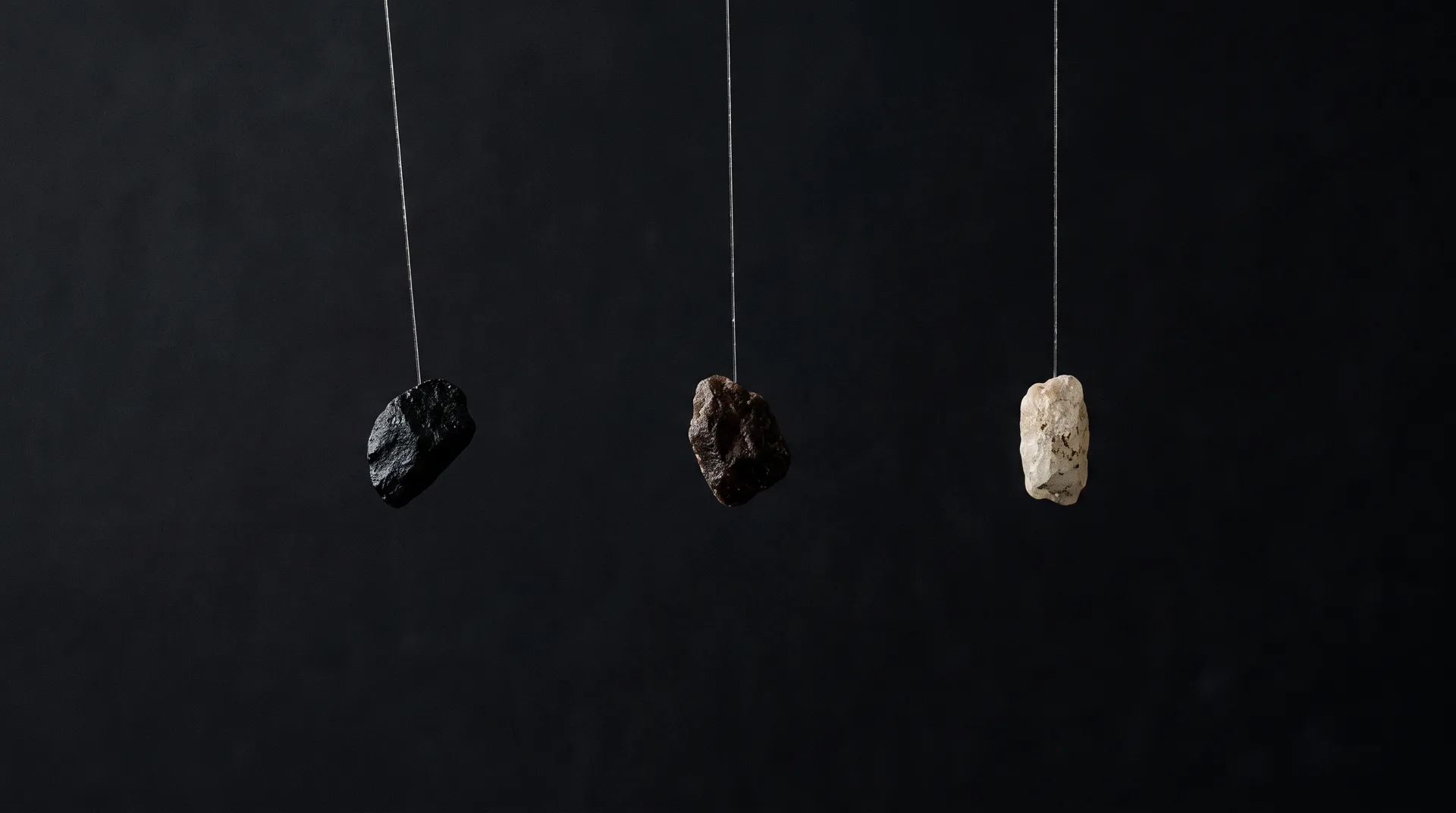 Three mineral grains hanging on fine threads against a dark background, a black grain pulled well aside, a brown one slightly, and a pale one hanging straight.