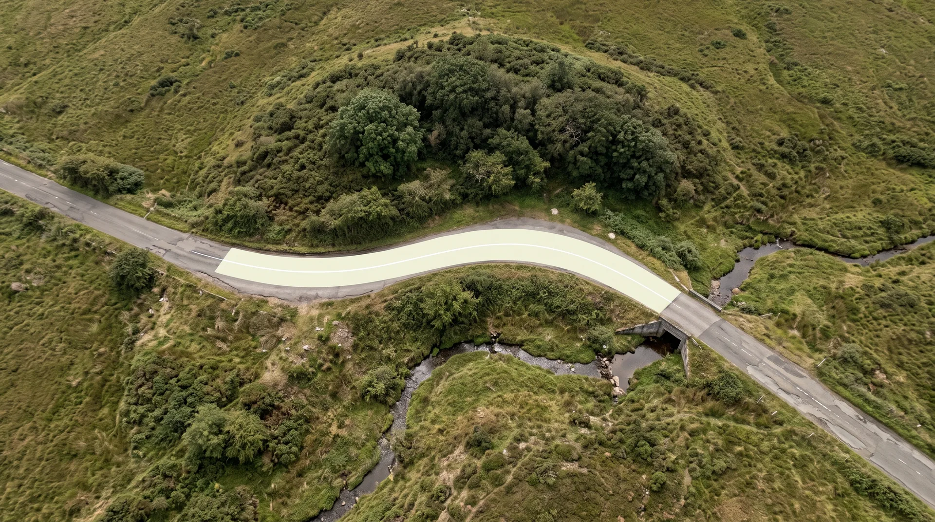 Aerial view looking straight down on an empty country road, one stretch newly marked with a pale painted lane, still curving around a wooded hill and a stream.